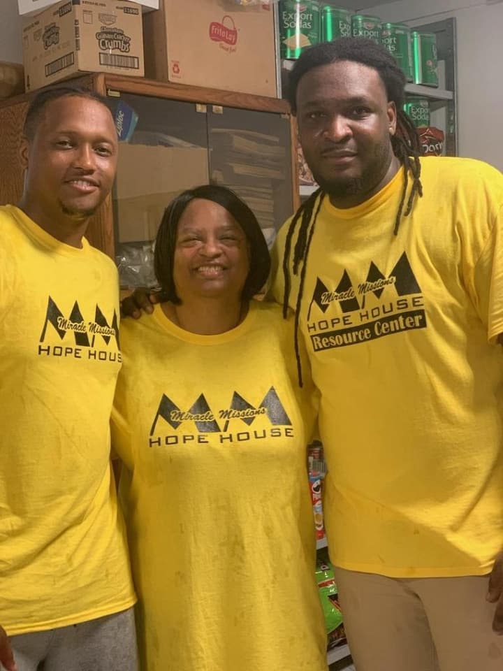 Three volunteers in yellow Miracle Missions Hope House Resource Center T-shirts smile together in the pantry storeroom.