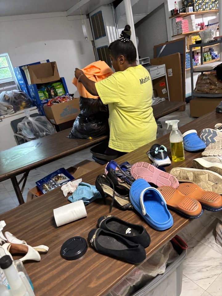 A volunteer in a yellow Miracle Missions Hope House T-shirt sorts donated shoes and clothing on a long table.
