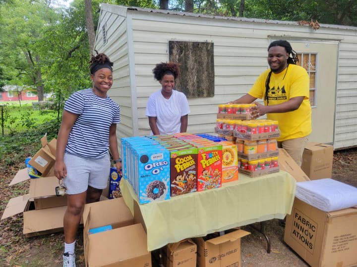 Miracle Mission volunteers smiling behind a table of donated cereal and peanut butter at an outdoor food distribution.